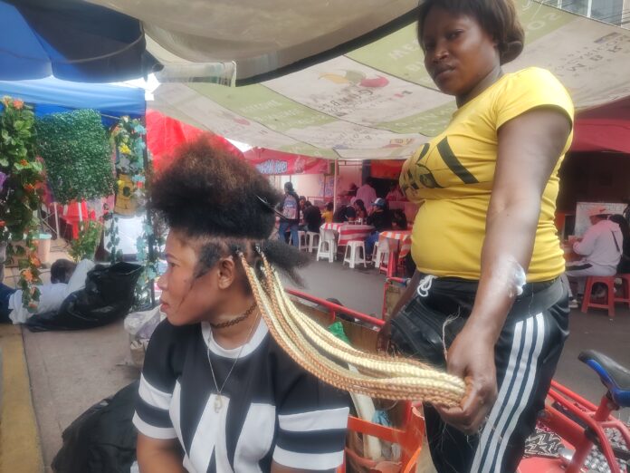 Theonilde y su clienta mientras trenza su cabello en el tianguis. Foto: Claudia Martínez